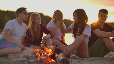 Two-boys-and-three-girls-are-sitting-around-bonfire-on-the-beach-with-beer.-They-are-talking-to-each-other-at-sunset-and-enjoying-the-summer-evening-on-the-river-coast.